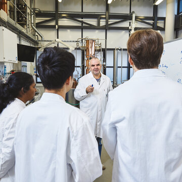 Lecture using the whiteboard in the laboratory A lecturer in a white lab coat stands in front of a whiteboard covered in handwritten notes and sketches, explaining the subject matter. Three students, also in white lab coats, stand with their backs to the camera, listening attentively. A large copper installation is set up in the background.