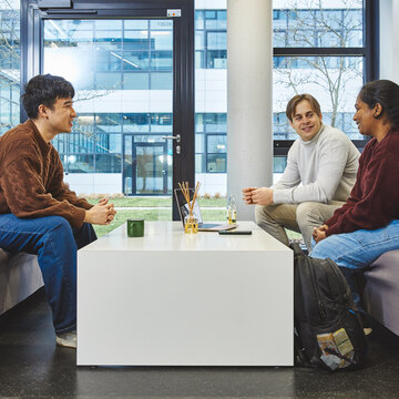 Exchange students on campus – Three students are sitting comfortably on sofas around a table in the foyer, chatting Three students in casual clothes are sitting opposite each other on two grey sofas in a bright, modern university building. On the white table between them are a laptop and some drinks. They are chatting and smiling.