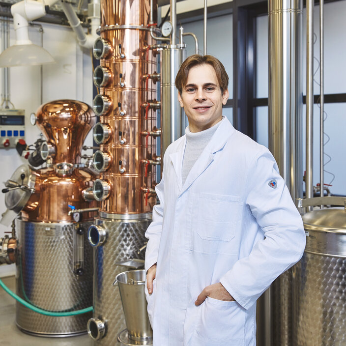 Student in front of an industrial laboratory unit – A smiling student in a lab coat is standing in front of a large distillation unit made of copper and stainless steel A young student in a white lab coat stands smiling in a laboratory, his hands in his coat pockets. Directly behind him stands a large, complex industrial plant made of gleaming copper and stainless steel.