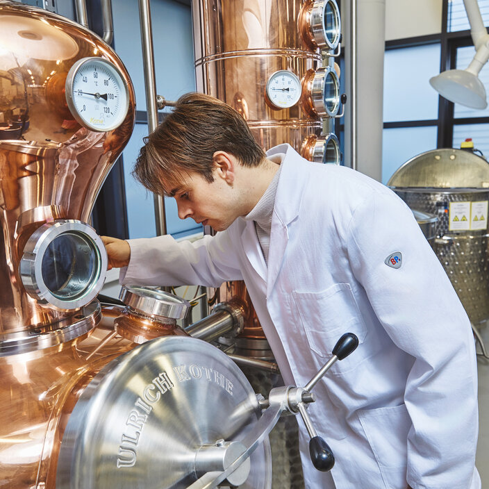 Practical work at the copper plant – A student in a white lab coat operates the valve on a large copper distillation plant A young student in a white lab coat is standing in profile in front of a large, gleaming copper apparatus. With one hand, he is grasping a black lever on a stainless-steel flap of the apparatus and is intently examining the machinery.