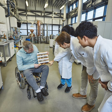 Analysis of soil samples – A lecturer in a wheelchair shows three students in lab coats a rack containing various soil and material samples In a brightly lit laboratory, a man in a wheelchair is sitting with a wooden display stand holding lots of small test tubes on his lap. Three students in white lab coats are leaning down towards him curiously, examining the samples.