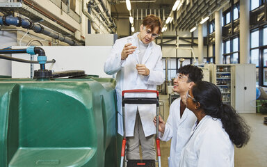 Water or algae samples at the bioreactor – A student is standing on a small ladder next to a large green tank, holding a beaker, whilst two others look on A student wearing a lab coat and safety goggles is standing on a small step in front of a large, green plastic tank. He is holding an empty beaker and talking to a male and a female student in lab coats, who are standing next to the tank and looking up at him.
