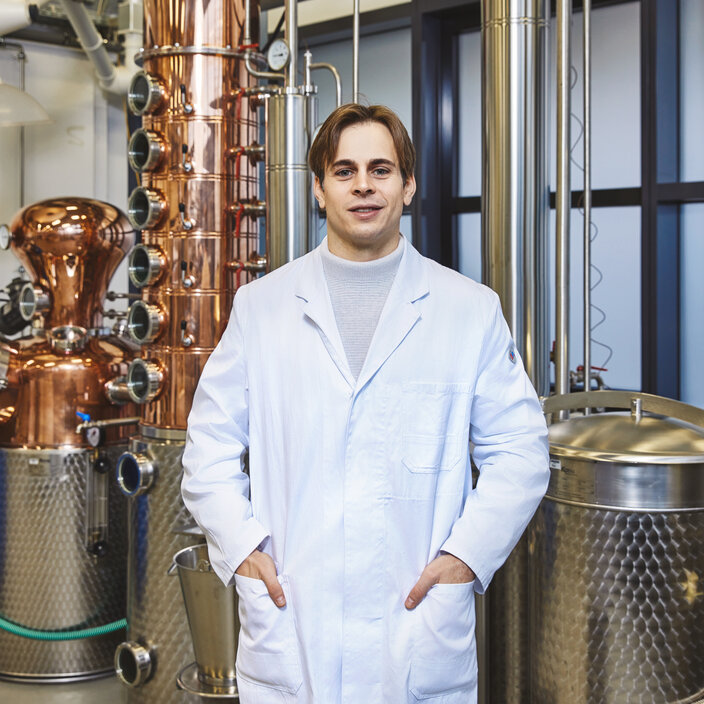 Practical learning in process engineering – a student in a lab coat stands in front of a state-of-the-art distillation unit made of copper and stainless steel A young student in a white lab coat stands smiling, with his hands in his pockets, in a brightly lit laboratory. Directly behind him is an imposing industrial installation made of gleaming copper and stainless steel.