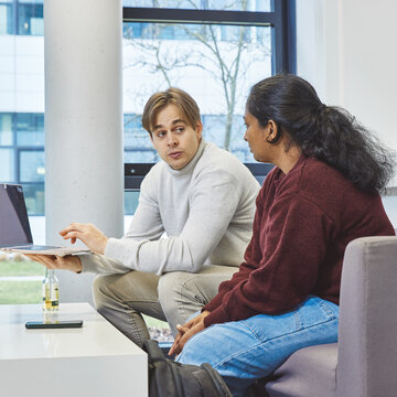 Working together on a project on a laptop – Two students are sitting on a sofa; one student is showing his fellow student something on a laptop he is holding up A male student and a female student are sitting next to each other on a grey sofa in a bright room with a window front. The male student is holding an open laptop in one hand and pointing at the screen with the other, whilst the female student looks at the display with interest.