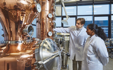 Students at a distillation plant A male student and a female student, both wearing white lab coats, are standing in a brightly lit laboratory in front of a large, shiny copper distillation apparatus. The male student is pointing to one of the round measuring instruments on the apparatus, whilst the female student watches intently.