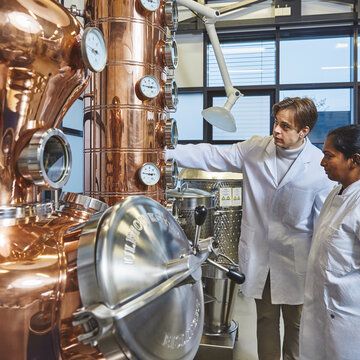 Students at a distillation plant A male student and a female student, both wearing white lab coats, are standing in a brightly lit laboratory in front of a large, shiny copper distillation apparatus. The male student is pointing to one of the round measuring instruments on the apparatus, whilst the female student watches intently.